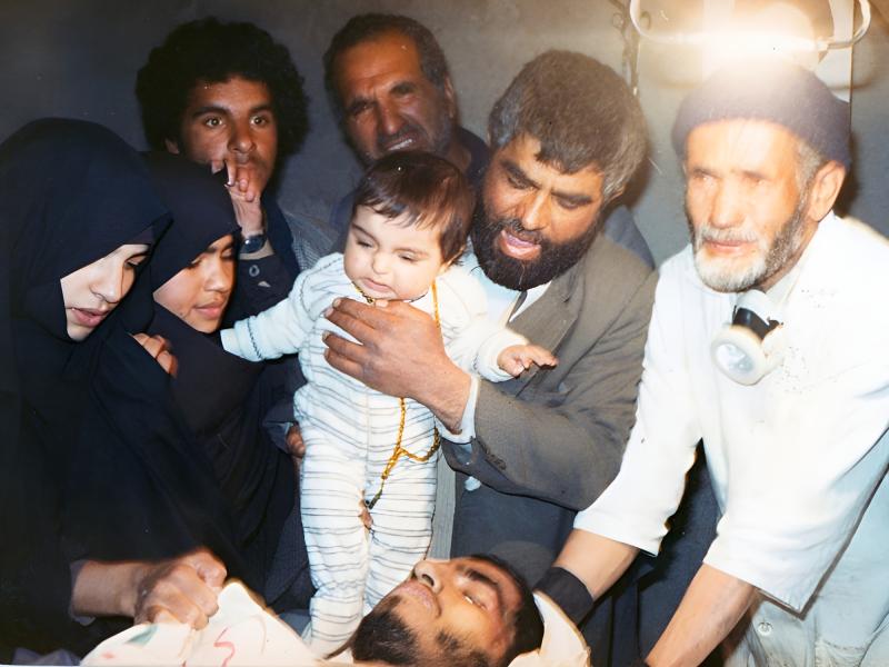 پدر شهید جهانگیر در حالی که نوهاش را در آغوش گرفته بر بالین پسرشهیدش آمده